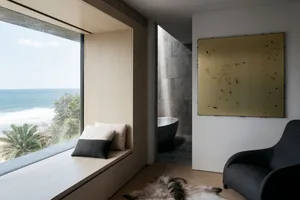 A modern residential interior featuring a window with a view of the ocean, a seating area with cushions, a large artwork, and