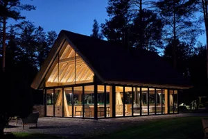 A modern residential building illuminated at night, featuring large glass windows and wooden structural elements.