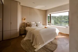 A modern bedroom with white cushions on the bed, a large window with a view of greenery, and a minimalist design.