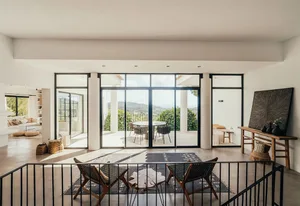 Modern minimalist living room with large glass windows and concrete floor, featuring natural-fiber seating and an outdoor vie