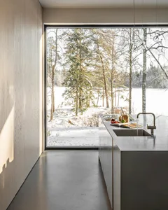 Modern kitchen with minimalist design featuring concrete countertops and floor, surrounded by a snowy forest view through lar