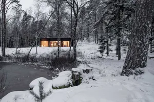 Modern residential cabin surrounded by snow-covered trees and a frozen pond at dusk, illuminated from within.