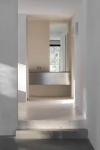 A modern residential corridor with light wooden flooring, concrete walls, a metal washbasin, and a view through a window.