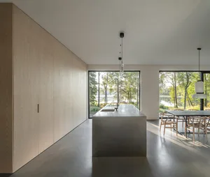 Modern residential interior featuring wooden cabinets, a concrete island, and large windows overlooking a forested area.