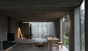 A minimalist interior space with concrete walls and floor, featuring a table, chairs, and a bed.
