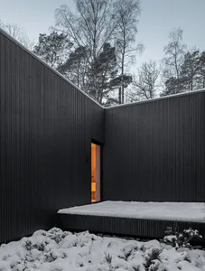 Modern house with black metal exterior and orange light visible from a window, surrounded by snow-covered landscape and trees