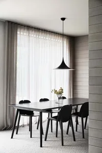 A minimalist dining room featuring a long wooden table with black chairs, large windows with sheer curtains, and a black pend