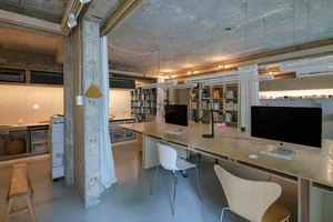 A minimalist office space with concrete walls, a long wooden desk, and white chairs.