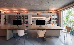 A minimalist office space featuring a concrete workstation with two computers, chairs, and bookshelves.