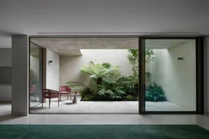 A modern living space featuring glass and concrete materials, with comfortable seating and lush greenery.