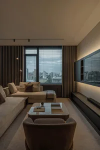 A modern minimalist living room with a beige sofa, armchair, coffee table with books, and large windows with blinds overlooki