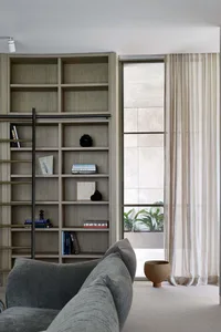 Contemporary residential library featuring wooden shelves and a gray sofa, with a large window and sheer curtains.