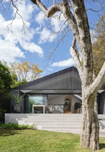 A modern house with a metal-clad exterior, featuring large glass windows and a sloping roof surrounded by greenery.