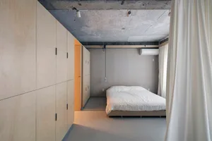 A minimalist bedroom with a concrete floor and walls, featuring a plain white bed with a simple wooden frame.