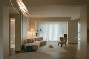 A modern minimalist living room with neutral-colored furniture, a large window with white curtains, and a clean, uncluttered