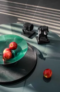 Three green glass decorative plates with ridges, containing three red apples, and two black ceramic statuettes of dogs on a g