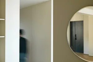 A minimalist interior featuring a person in a suit walking through a hallway with light-colored walls and flooring.