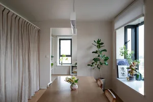Modern living room featuring a wooden dining table adorned with flowers, surrounded by plants and glass window, with a fabric
