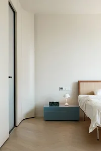 A modern, minimalist bedroom featuring a bed with a white fabric headboard and a nightstand with a blue drawer and a white la