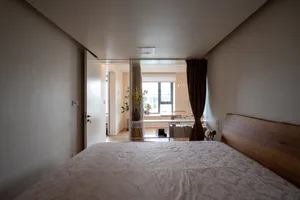 A minimalist bedroom with a wooden headboard, beige fabric bedding, and subtle patterned bedding spread across a bed, with a