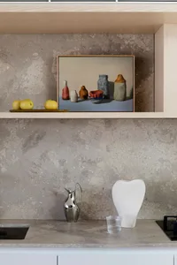 A modern kitchen counter with a grey concrete finish, featuring a metal pitcher, a white vase, and an artwork of vases and ap