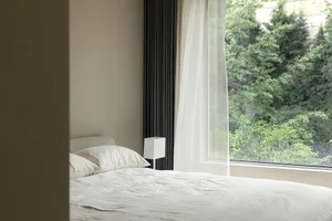 A minimalist bedroom with a bed featuring white textiles and natural light streaming through a large window with a sheer curt