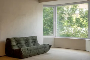Minimalist living room featuring a green upholstered sofa in front of a large window with a view of trees.