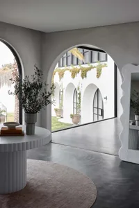 A modern architectural courtyard with concrete flooring, arches, and greenery, featuring a round table with a vase and books.