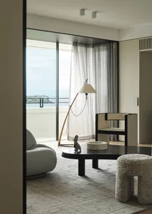 A modern living room with soft textiles, featuring a round coffee table, a floor lamp, and a view of the ocean through floor-