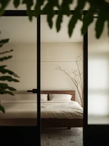 Minimalist bedroom with a wooden bed frame and white bedding, viewed through a glass door with green foliage outside.