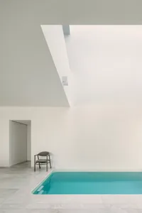 A minimalist indoor swimming pool with a white marble floor, surrounded by white walls and a ceiling with a skylight.