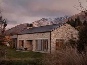 A rustic stone residential exterior with a metal roof and a stone chimney, set against a mountainous backdrop.