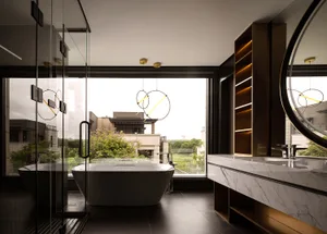 Modern bathroom with marble tub, glass shower, and large windows overlooking greenery.