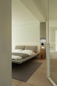 Modern, minimalist bedroom with a floating platform bed, grey headboard, and a wooden stool with a lamp on top.