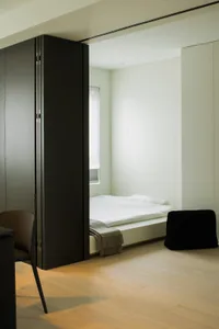 Minimalist bedroom showcasing a white bed placed on a wooden floor, featuring a black chair and a black cushion.
