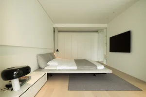 A minimalist modern bedroom with a flat-pack bed, white walls, and a television mounted on the wall.