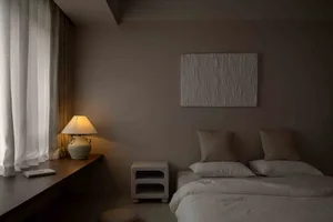 Minimalist bedroom with neutral tones, featuring a bed, nightstand, lamp, and artwork