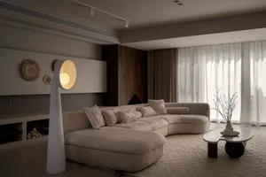 A modern living room with a curved sofa, floor lamp, and large windows with white curtains.