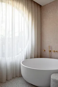 A modern minimalist bathroom with a white freestanding tub, blush pink mosaic tiles, and a sheer curtain.