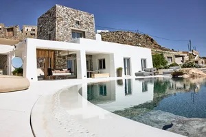 A minimalist residential exterior featuring white plaster and stone materials, situated on a hillside with an infinity pool.