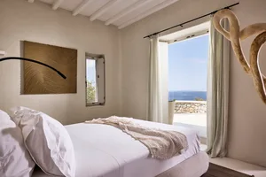 A minimalist bedroom with neutral tones, featuring a white bed, light-colored curtains, and a view of the ocean through the w
