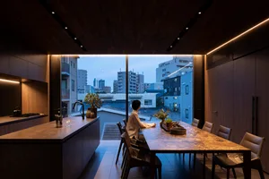A minimalist kitchen with a marble countertop, modern seating, and a view of urban buildings through a large window.