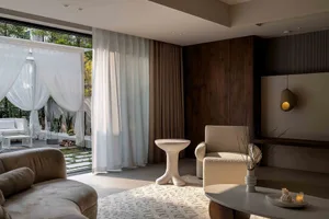 A modern living room featuring a white sofa, armchair, and side table with a glass top, set against a backdrop of large windo