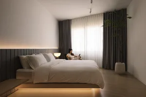 A minimalist bedroom featuring a bed with a textured wooden headboard and floor, white bedding, and a potted plant in the cor