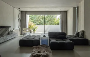A modern minimalist living room with a concrete floor, large windows, and a large black modular sofa.