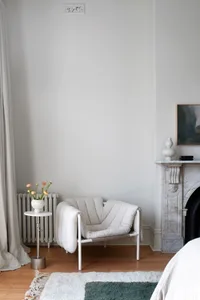 A minimalist residential interior with a white fabric armchair, marble fireplace mantel, and a green and white rug on a woode