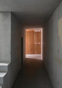 A narrow corridor with concrete walls and wooden paneling, featuring a window at the end letting in natural light.