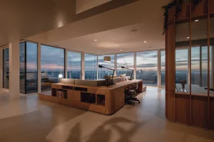 Modern living room with wooden furniture, large windows, and a view of a cityscape during sunset.