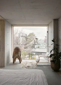 A minimalist bedroom with concrete walls, a white bed, a white chair, and a plant in a large window with a city view.