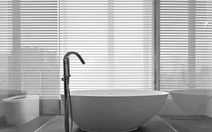 Modern bathroom with a white ceramic freestanding bathtub in front of a window with white blinds.
